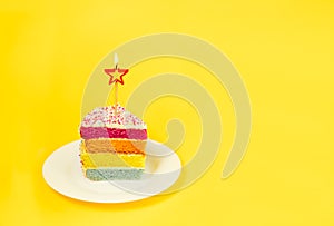 Slice of Rainbow cake with birning candle in the shape of star on white round plate isolated on bright yellow background. Happy