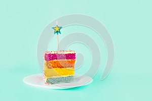 Slice of Rainbow cake with birning candle in the shape of star on white round plate isolated on turquoise background. Happy