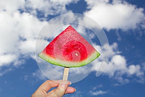 A slice of watermelon on a wooden stick in his hand against the background of the sky with clouds.