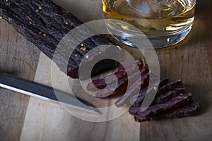 Sliced beef biltong on wooden board with knife and a glass of whisky