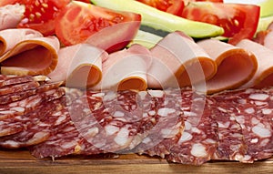 Sliced dry sausages and meat products, cured meat, bacon, with fresh cucumber, tomatoes slices on a wood board