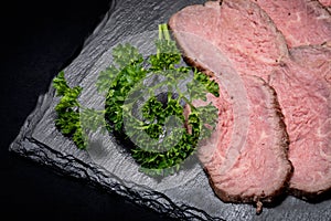 Sliced Grass Fed Juicy Corn Roast Beef garnished with Fresh Curly Parsley on black natural stone