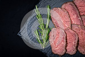 Sliced Grass Fed juicy Corn Roast Beef garnished with Rosemary Fresh Herb on natural black stone