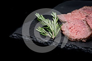 Sliced Grass Fed juicy Corn Roast Beef garnished with Rosemary Fresh Herb on natural black stone