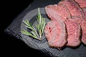 Sliced Grass Fed juicy Corn Roast Beef garnished with Rosemary Fresh Herb on natural black stone