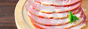Sliced ham with fresh green lettuce leaves on a round cutting board. Meat products on a brown wooden table.. Banner