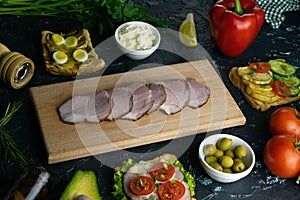 Sliced ham on a wooden board, around the ham, green onions, tomato, souce, olives, lemon, spice, dill and parsley