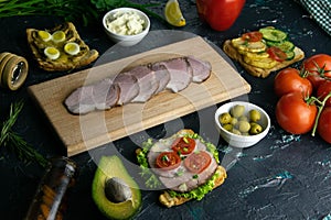 Sliced ham on a wooden board, around the ham, green onions, tomato, souce, olives, lemon, spice, dill and parsley