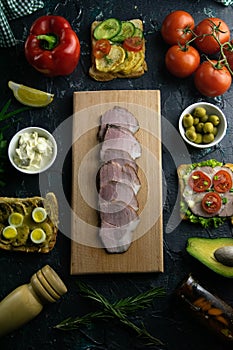 Sliced ham on a wooden board, around the ham, green onions, tomato, souce, olives, lemon, spice, dill and parsley