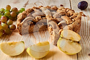 Sliced homemade american apple pie on brown wooden table with raisins and fresh fruits - apples, plums and grapes