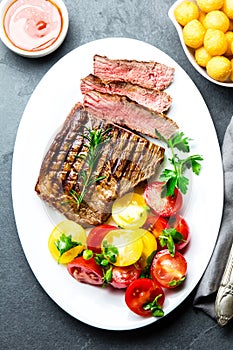 Sliced medium rare grilled beef steak served on white plate with tomato salad and potatoes balls. Barbecue, bbq meat