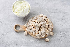 Sliced mini champignon mushrooms on a wooden board and a bowl of chopped onions on a gray textured background. Cooking