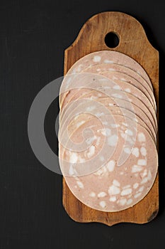 Sliced Mortadella Bologna Meat on a rustic wooden board over black background, top view. Flat lay, from above, overhead. Copy
