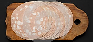 Sliced Mortadella Bologna Meat on a rustic wooden board over black surface, top view. Flat lay, from above, overhead