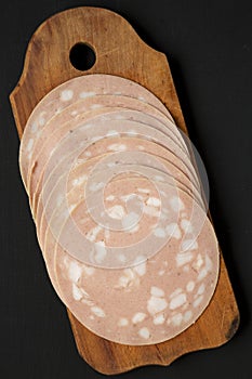 Sliced Mortadella Bologna Meat on a rustic wooden board over black surface, top view. Flat lay, from above, overhead. Close-up