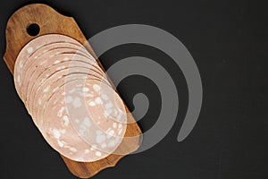 Sliced Mortadella Bologna Meat on a rustic wooden board over black surface, top view. Flat lay, from above, overhead. Space for