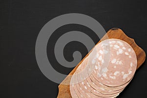 Sliced Mortadella Bologna Meat on a rustic wooden board over black surface, top view. Flat lay, from above, overhead. Space for