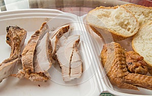 Sliced pieces of cold baked pork in a plastic container, with slices of wheat bread and fried chicken wing, an