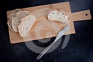 Sliced pieces go ciabatta bread on the wooden cutting board with butter on it and buttered knife next to it.