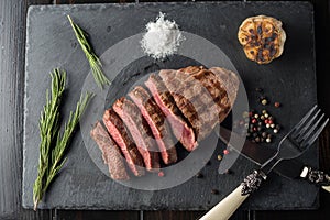 Sliced prime ribeye steak on black stone plate. Medium degree of steak doneness. With rosemary and peppers. Fork and knife.