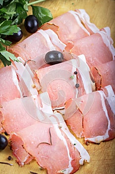 Sliced prosciutto ham with black olives and spices on wooden background