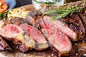 Sliced rare grilled or barbecued tomahawk beef steak on a griddle with fresh rosemary and tomato in a close up view.