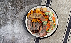 Sliced roast beef with baked potatoes and tomato and cucumber salad on a round plate on a dark background