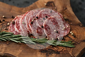 Sliced salame milano sausage on olive wood board with rosemary and pepper
