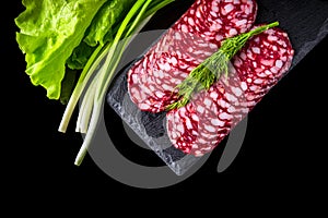 Sliced salami with dill and salad greens on a slate serving Board. Black isolated background