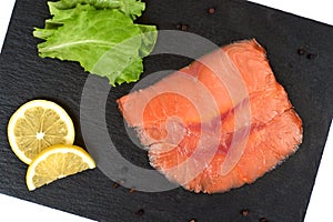 Sliced salmon fillet on the black shale board, with lemon and salad. On white background