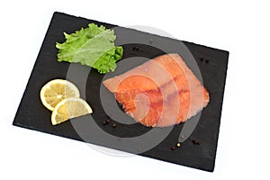 Sliced salmon fillet on the black shale board, with lemon and salad. On white background