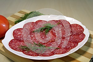 Sliced sausage on a white plate with dill on a wooden board.