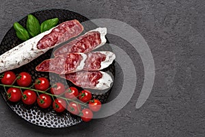 Sliced thin catalan fuet sausage and bunch of red cherry tomatoes on a black plate over dark rough surface. Traditional spanish