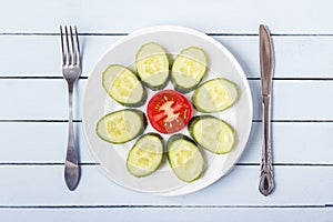 Sliced tomato and cucumber for healthy breakfast on white plate. Fork and knife on wooden table. Top view. Copy space