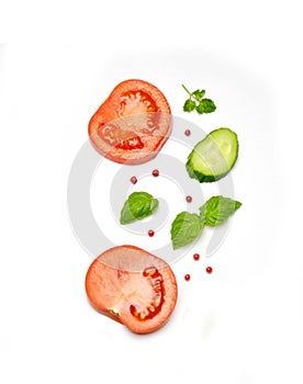 Sliced tomato and cucumber next to Basil leaves and pepper on a white