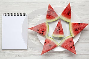 Sliced watermelon on white plate with blank notepad over white wooden background, top view.