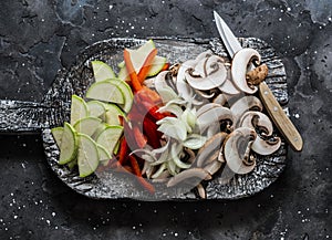Sliced zucchini, mushrooms, sweet paprika, onions on a rustic chopping board on a dark background, top view. Raw food ingredients
