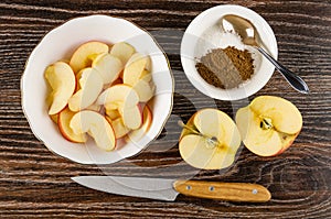 Slices of apples in bowl, spoon in bowl with sugar and ground cinnamon, half of apple, knife on wooden table. Top view