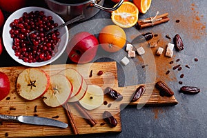 Slices of apples, cinnamon and dates on a board, next to a plate with cranberries and a saucepan. Indoors.