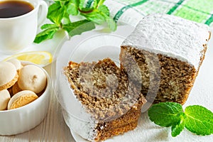 Slices of biscuit cake, a ÃÂup of tea with lemon, small cookies and mint leaves on a white wooden table