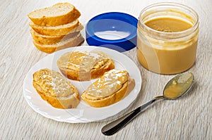 Slices of bread, cap, jar with peanut butter, sandwiches with peanut paste in plate, spoon on wooden table