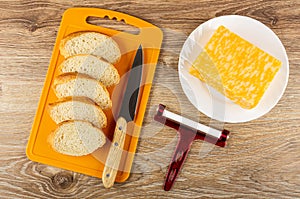Slices of bread, knife on cutting board, piece of marble cheese in plate, cheese cutter on table. Top view