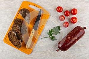 Slices of bread, knife on cutting board, tomato, parsley, smoked sausage in polyethylene pack on table. Top view
