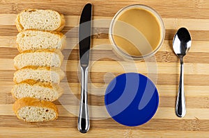 Slices of bread, knife, jar with peanut butter, blue cap, spoon on striped table. Top view