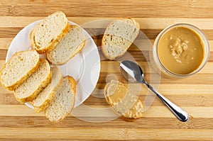 Slices of bread in plate, sandwich with peanut paste, spoon, jar with peanut butter on bamboo table. Top view