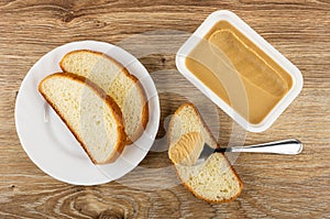 Slices of bread in plate, spoon with peanut butter on slice of bread, box with peanut paste on table. Top view