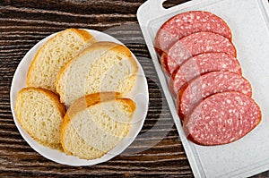 Slices of bread in plate on wooden table, slices of cervelat on cutting board. Top view