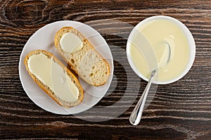 Slices of bun poured condensed milk in plate, bowl with condensed milk on wooden table. Top view