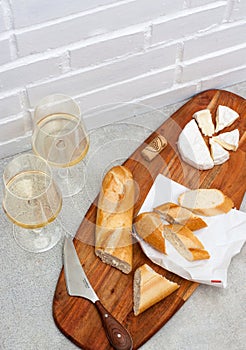 Slices of camembert cheese, baguette and two glasses of white wine on wooden board. White brick wall as background