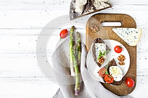 Slices of dark bread with blue cheese, eggs, tomatoes on wooden cutting board decorated with asparagus. Flat lay, top view